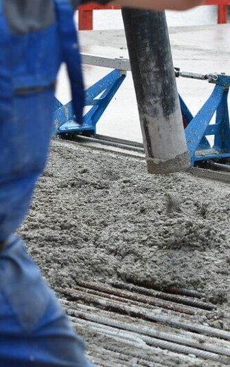 Ein Arbeiter in blauer Kleidung steht im Vordergrund, während aus einem dicken Schlauch frischer Beton auf eine Baustelle gegossen wird. Im Hintergrund ist eine blaue Abziehschiene zu sehen, die zur Glättung der Oberfläche dient. Unter dem Beton verlaufen mehrere gespannte Stahlseile oder -litzen, die für die Vorspannung im Betonbau verwendet werden. Die Szene zeigt einen typischen Arbeitsschritt beim Einbau von Spannbeton.

                                    